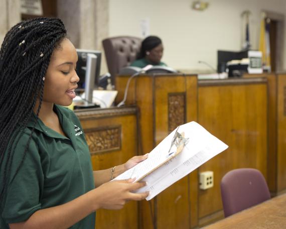 Youth Impact participant in green short reads from an agenda in a courtroom