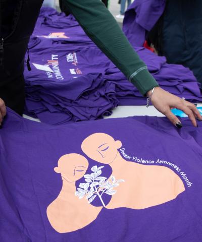 Woman holding out a purple shirt that reads "Domestic Violence Awareness Month" next to  representation of two women with a plant growing between them.