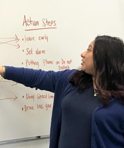 A woman points to a whiteboard with two columns labeled "Causes of Risky Driving" and "Action Steps," with arrows connecting examples listed on either side.