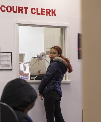 Woman in a winter jacket stands in front of an indoor booth below the words "Court Clerk" in bold red letters.