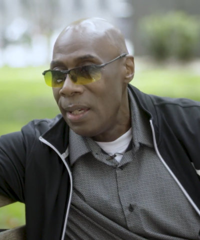 Middle-aged man with yellow sunglasses and a button-down shirt on a park bench during the day.