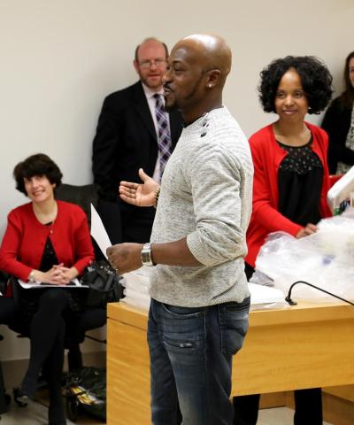 A man in sweater and jeans speaks in front of a group of people smiling