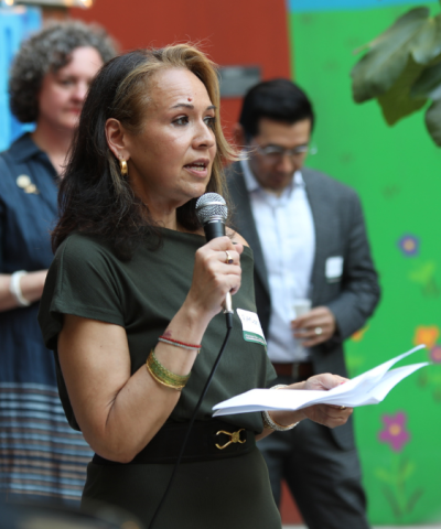 Rasmia Kirmani speaks holding a microphone in front of green floral mural in a community garden