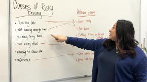 A woman points to a whiteboard with two columns labeled "Causes of Risky Driving" and "Action Steps," with arrows connecting examples listed on either side.