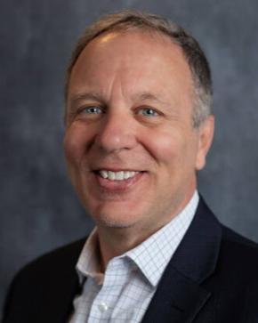 White man with traditional suit, no tie, smiles into the camera against a gray backdrop