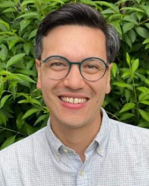 Asian man with glasses and a checkered white/blue shirt smiles into the camera against a background of green leaves