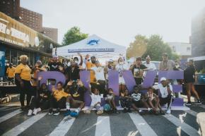 Large group of people wearing purple shirts pose in front of a purple "BLIVE" sign