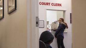 Woman in a winter jacket stands in front of an indoor booth below the words "Court Clerk" in bold red letters.