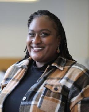 Black woman in brown and black plaid smiles into the camera lens against a light gray background